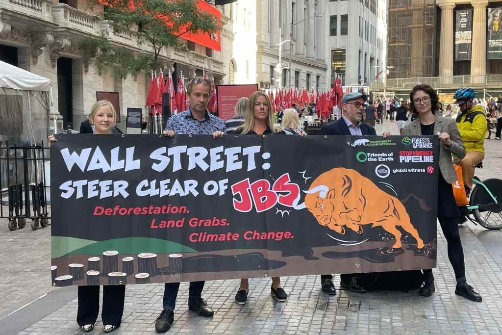 Five people stand on a city street holding a large protest banner that reads “Wall Street: Steer clear of JBS — Deforestation. Land Grabs. Climate Change.”