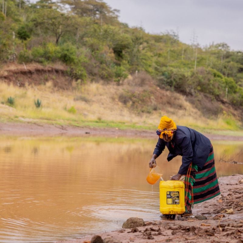 A woman is pouring water into a big yellow plastic bottle by a lake in Africa.