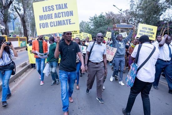 A group of people are participating in a street march holding signs saying 'Less talk more action for climate'